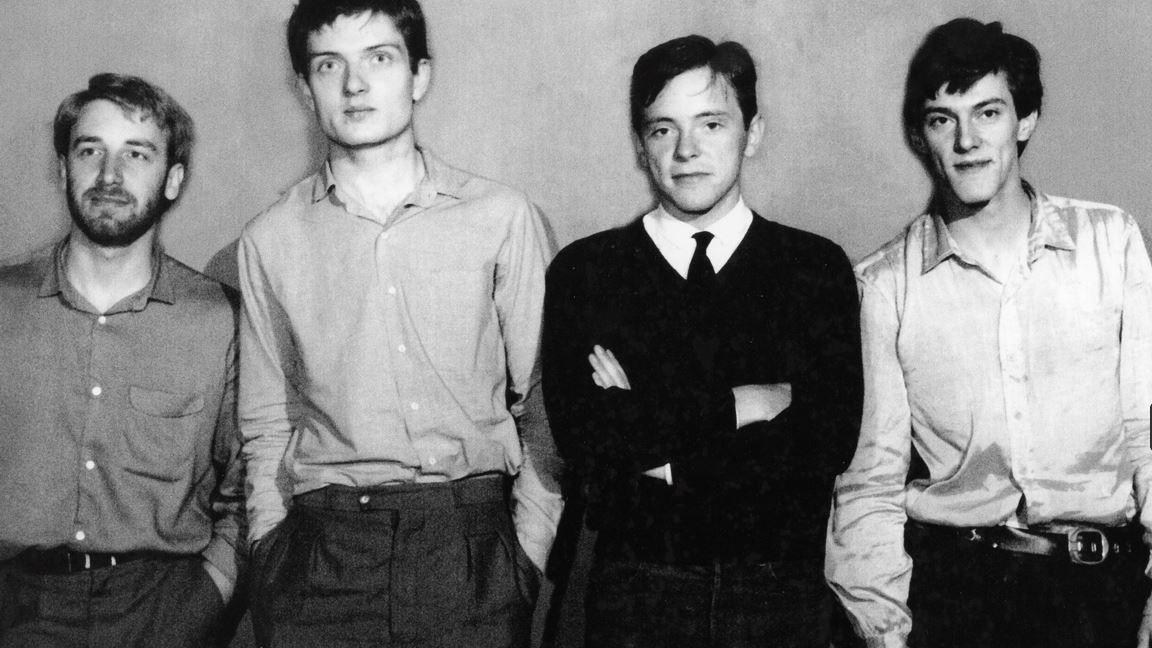 The four members of the band in a black and white snap from the 1970s. They are standing wearing shirts and trousers. Bernard Sumner is also wearing a black jumper and tie and has his arms folded. 