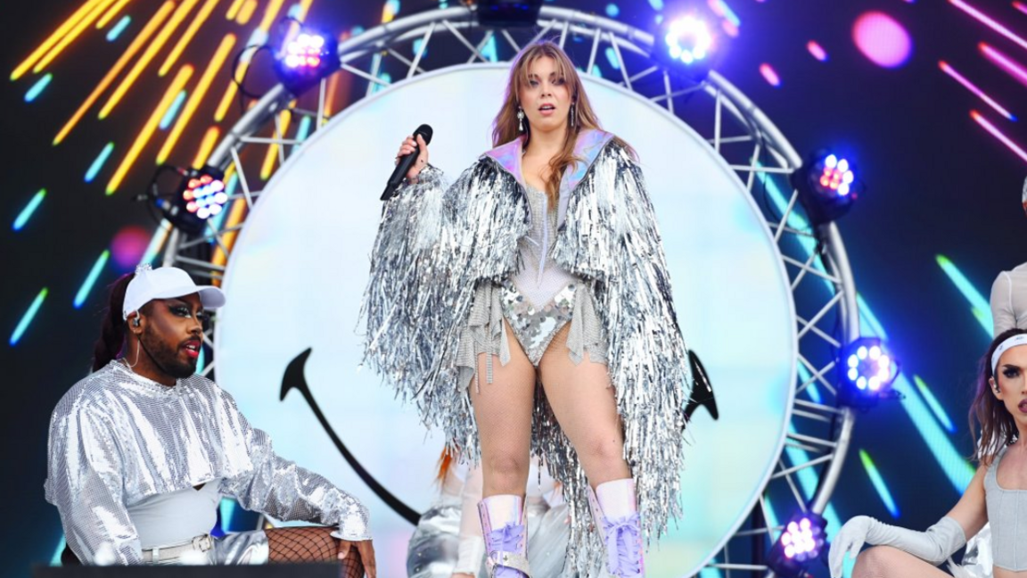 Becky Hill, wearing a silver sparkly jacket, and lilac boots performs on stage with two dancers at her feet