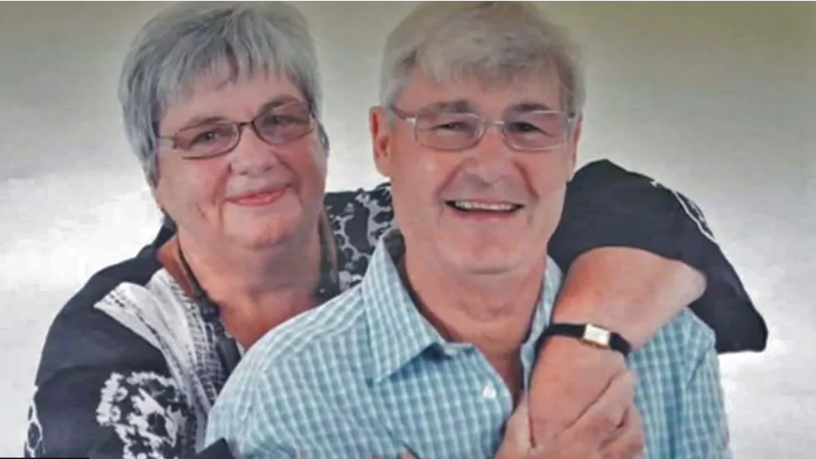 Picture of a woman hugging a man from behind. Both couple have grey hair, are wearing glasses and smiling. The man is wearing a blue and white checked shirt while his wife is wearing a black and white shirt, a black chunky beaded necklace and a square framed watch with black leather straps.