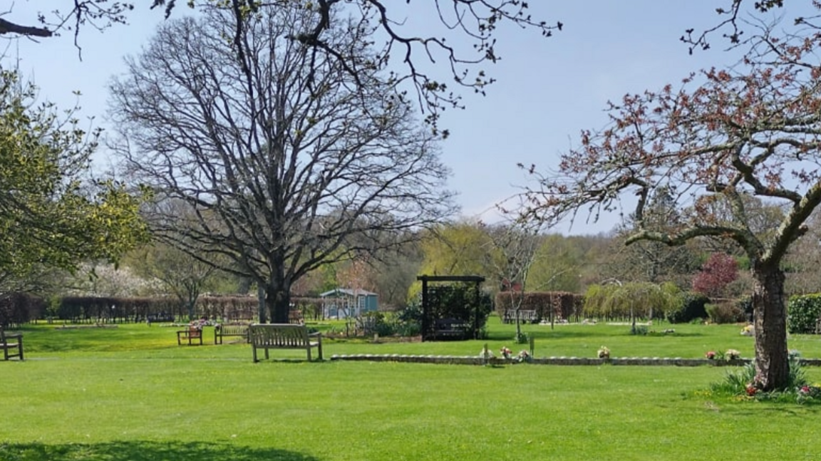 Isle of Wight Crematorium memorial gardens. It has several trees, an expanse of green grass and a number of wooden benches.