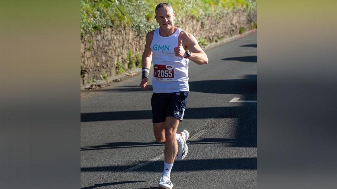 A man in running gear gives a thumbs up while running along a country road with a hedgerow by the side.