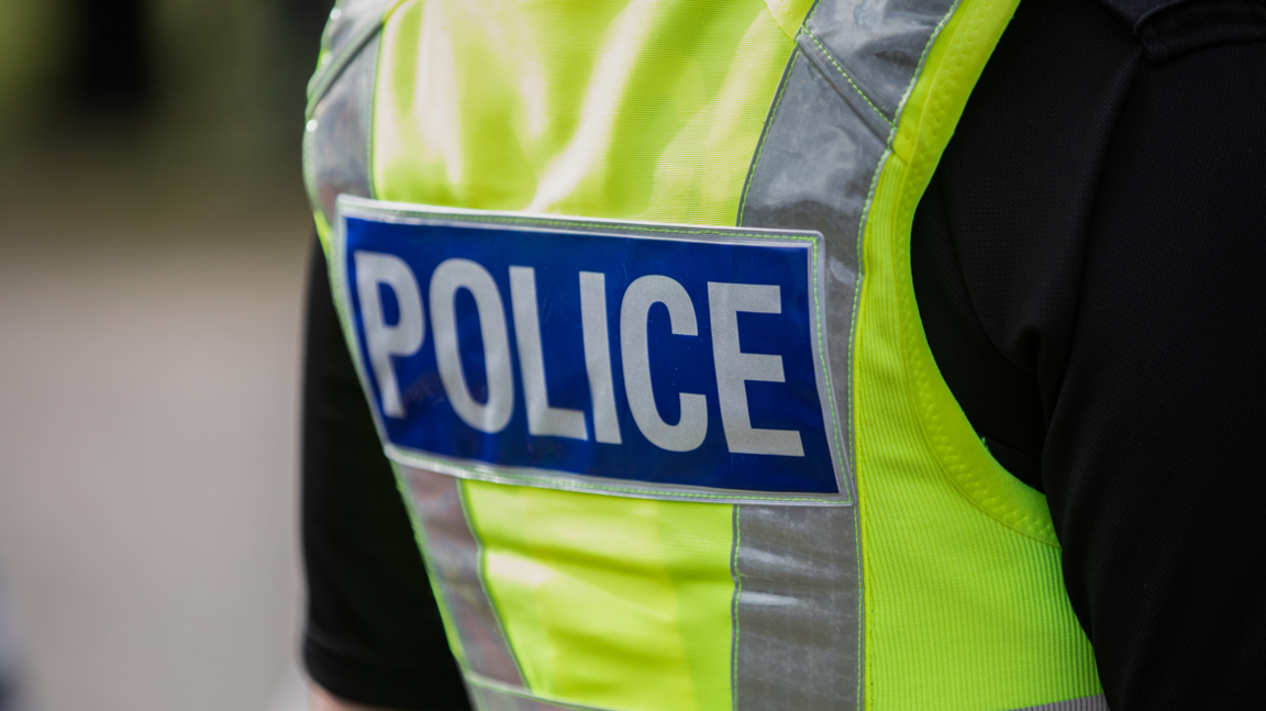 A close-up image of the back of a police officer in uniform, focusing on his high-viz yellow jacket which says Police on it in a blue rectangle. The background is blurred.