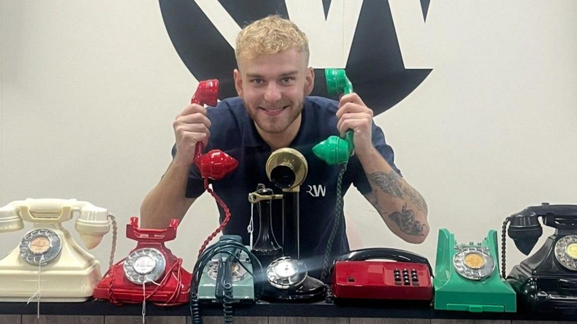 A man with blond hair is smiling at the camera and holding the receiver of a red phone in his right hand and of a green phone in his left hand. Seven phones are on the flat surface in front of him.