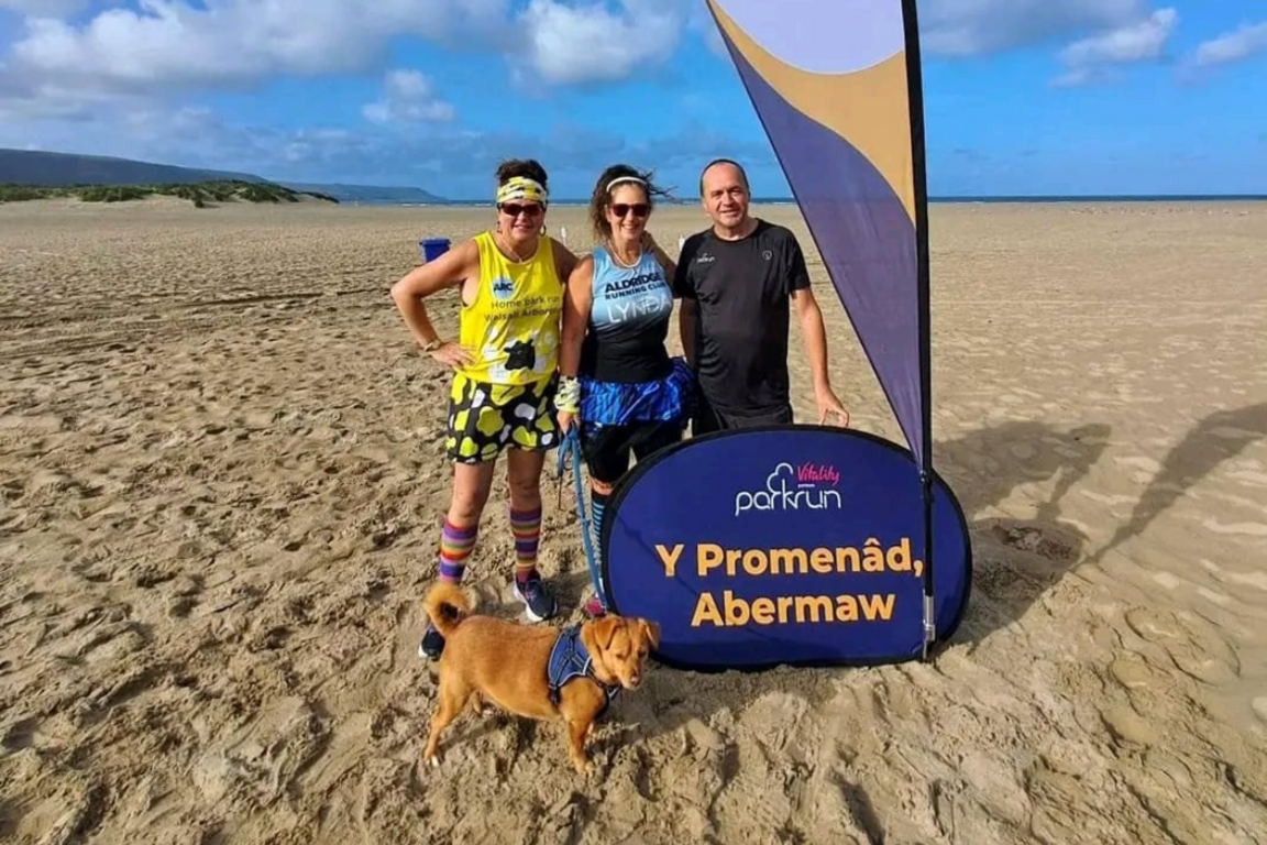 Two women and a man stand on a beach in running gear, with one of the women holding the lead to a small brown dog. They stand next to a purple Parkrun sign saying Y Promenâd, Abermaw