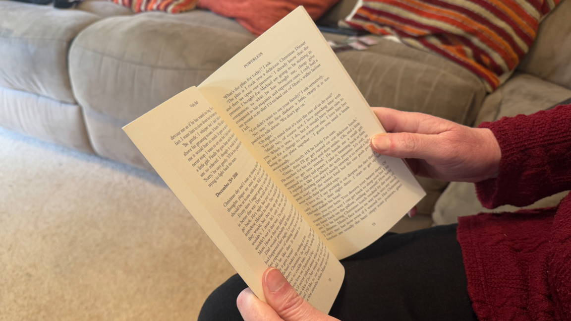 An open book being held in the hands of a woman who is out of shot. She is sat on a sofa inside.