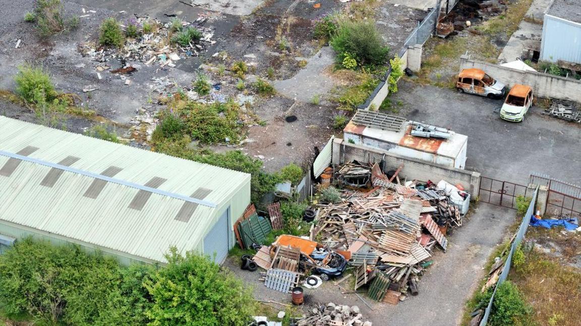 A overhead view of the disused industrial site. Piles of pallets are contained within a yard, while burnt out cars have been abandoned nearby.