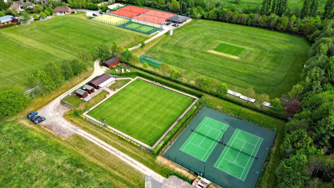 An aerial view of tennis courts at Ramsbury Tennis Club. Its shows several grass courts from above and some red clay-style courts in the distance.