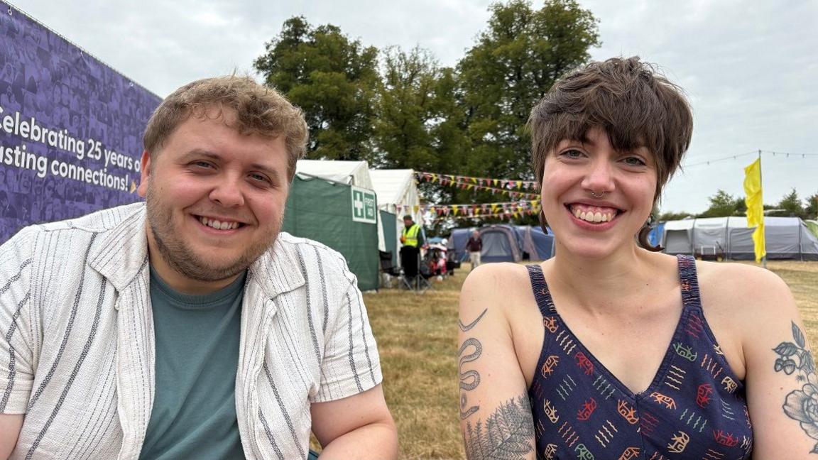 Eleanor Crame with medium-length dark hair wearing a blue top and tattoos, and Matthew Crame with short fair hair wearing a green T-shirt and white shirt. There is a large blue banner behind Matthew and marquees in the distance.