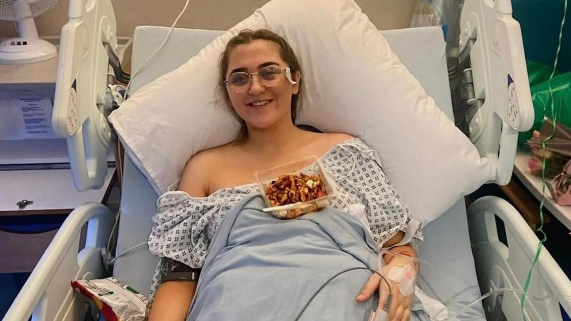 Ami is propped up in a hospital bed with a large white pillow behind her head and tubes coming out of her hand - there is a pot of pasta in front of her and she is smiling