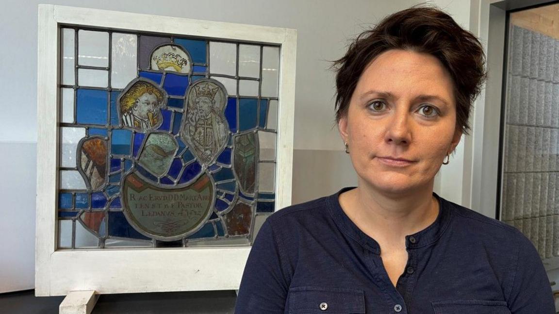 A woman with short dark hair, brown eyes and wearing a blue collarless shirt stands next to a panel of stained glass. It shows detailed faces and portions of blue and orange glass.