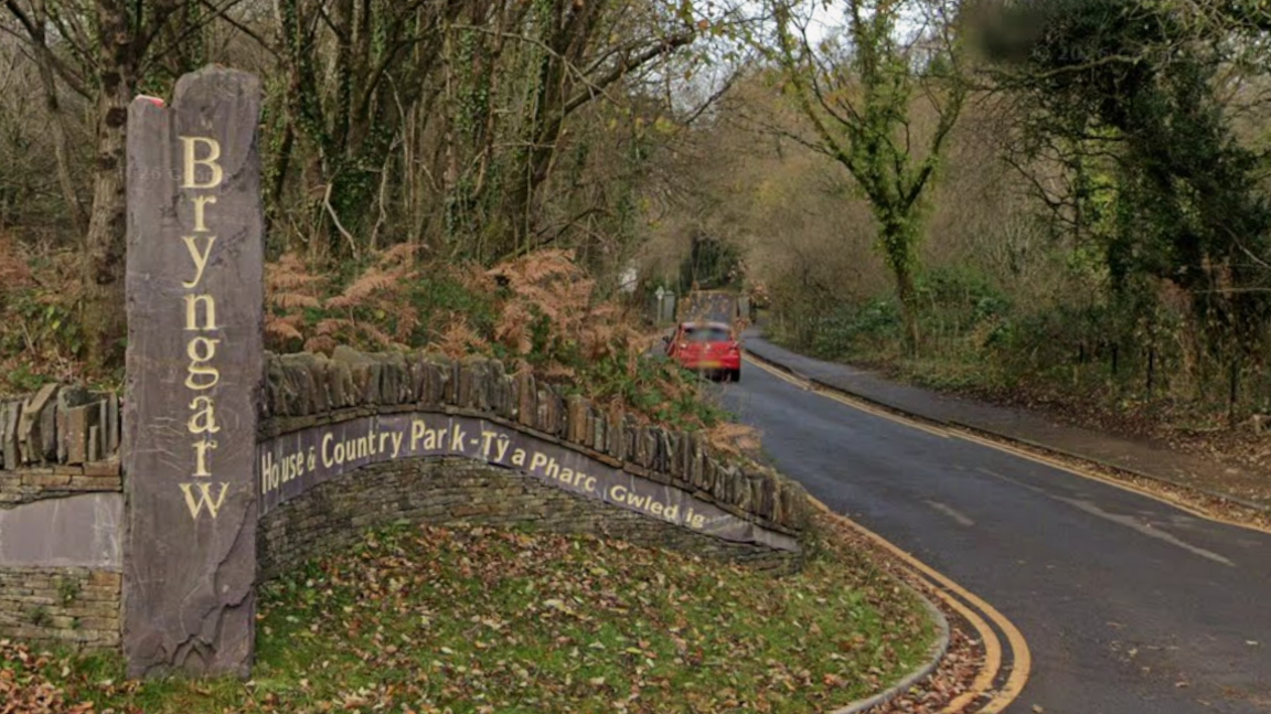 A tree-lined country road curves gently through a wooded landscape at the entrance to Bryngarw House and Country Park. In the foreground, a tall vertical stone marker carved with the word “Bryngarw” stands beside a curved stone wall bearing bilingual Welsh and English signage for the park. Brown ferns, ivy-covered trees and fallen leaves surround the entrance. A small red car is visible further down the road, which disappears into the wooded park beneath a canopy of bare branches.