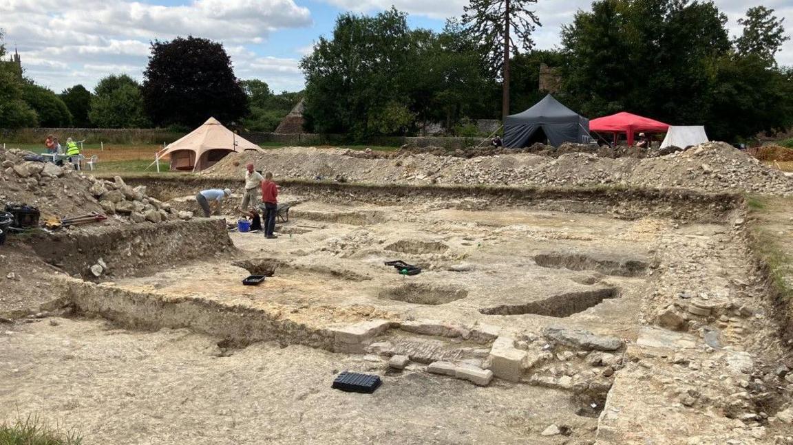 A large excavated area of chalk and stone revealing the base of stone walls. There are a number of pits and three people working in the area. In the background are a number of tents and gazebos.