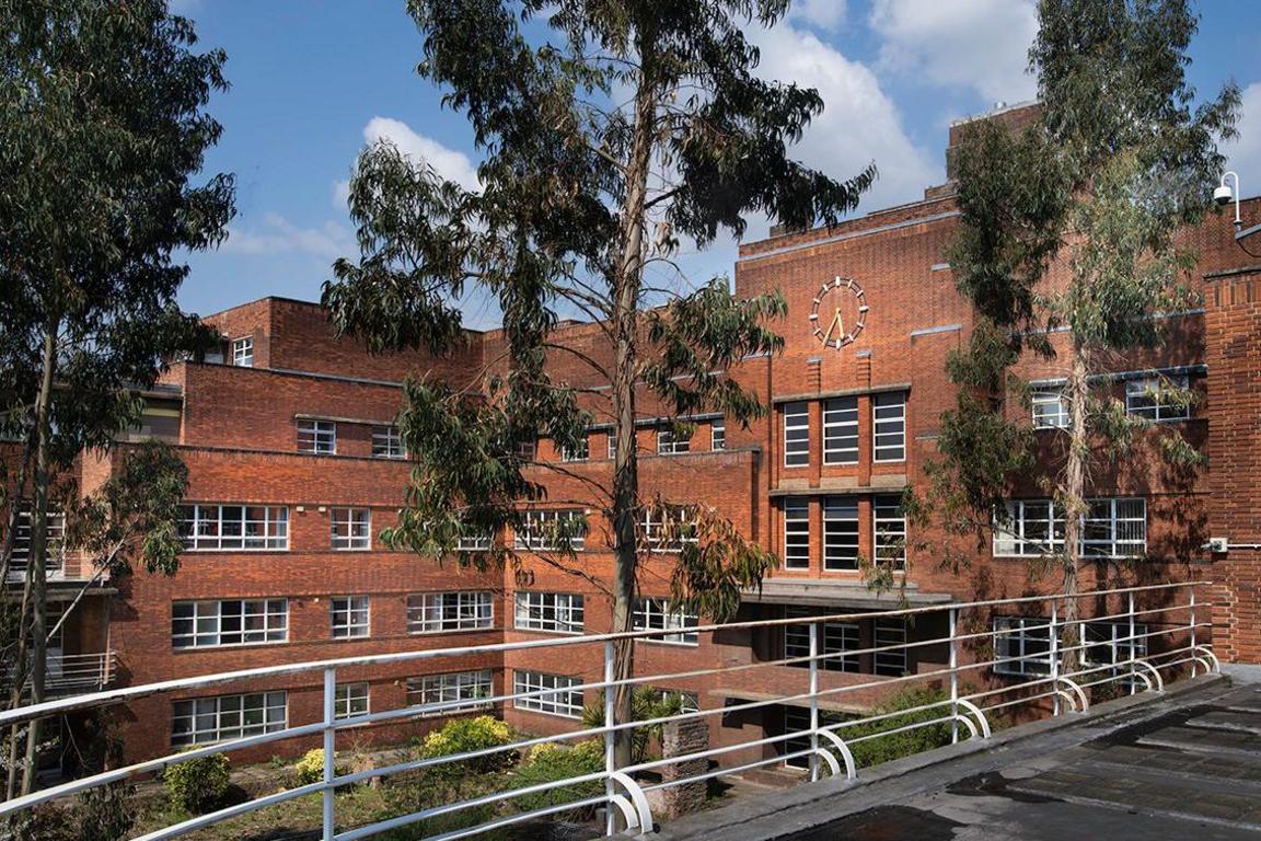 Taken from a balcony on the third floor of a building, overlooking the other two sides of the building (on the right and in front) and a courtyard. The balcony has a white railing, beyond which there are three tall trees with sparse green leaves. Behind these trees, the rest of the building is visible. It is a red-brick building with white-framed windows on each floor, and a clock at the top of the middle of the building. The courtyard below has a few bushes and green grass. The sky behind the building is blue, with a few clouds.