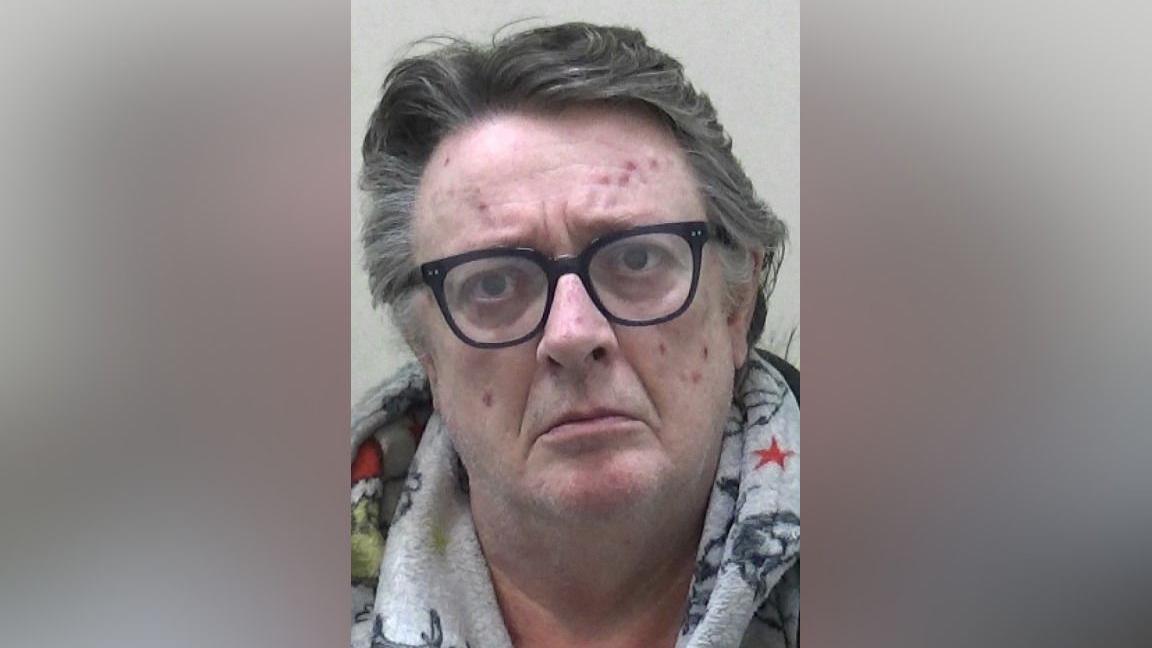 Mugshot of Storey. He is clean shaven with thick long grey hair swept back and various red spots and marks on his face. He is wearing black glasses and a grey hoody with colourful characters on it, possibly the Grinch.