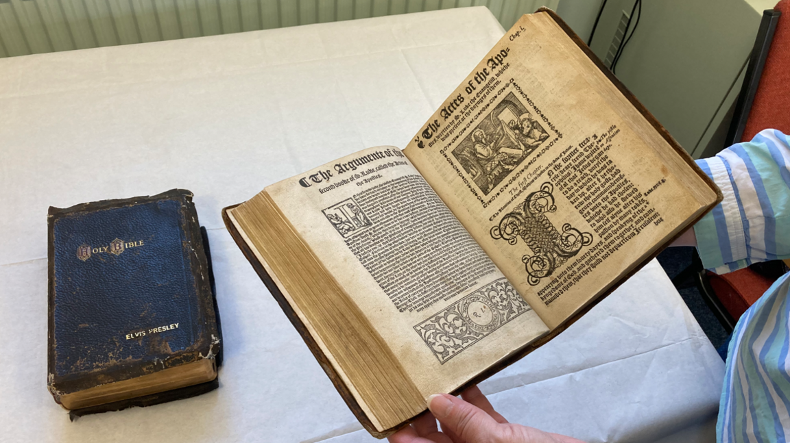 Elvis Presley's bible is rested on a table covered in a white cloth to the left of the image. An open bible is being held by female-looking hands.