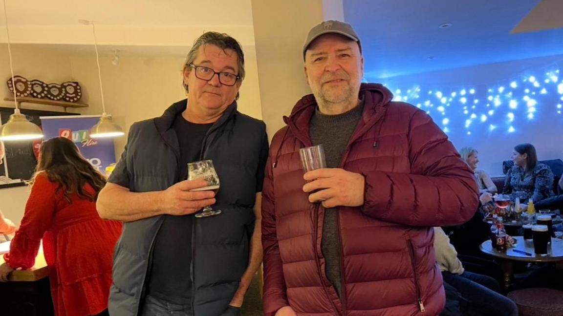 Two men holding glasses of beer. The man on the left is wearing a dark blue bomber jacket and a dark blue T-shirt. On the right is a man wearing a crimson jacket and a cap. They are inside a bar, with other people in the background.