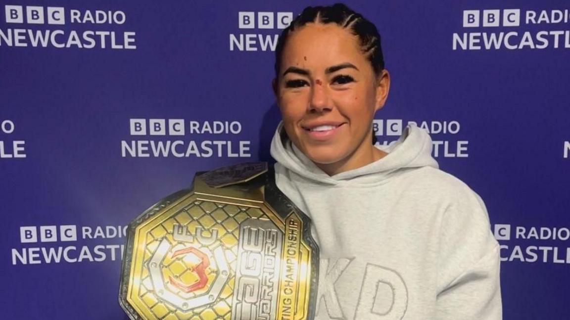 Kennedy Freeman, wearing a cream hoodie and holding her newly won World Championship belt, which is big and coloured gold, silver and steel, with the championship logo on it. She's standing in front of a screen with BBC Radio Newcastle written repeatedly all over it.