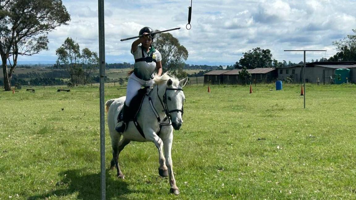 Khosi Dlomo rides on her horse Yogi and holds a lance aloft in her outstretched right arm as she attempts to strike at a circular target hanging from a pole