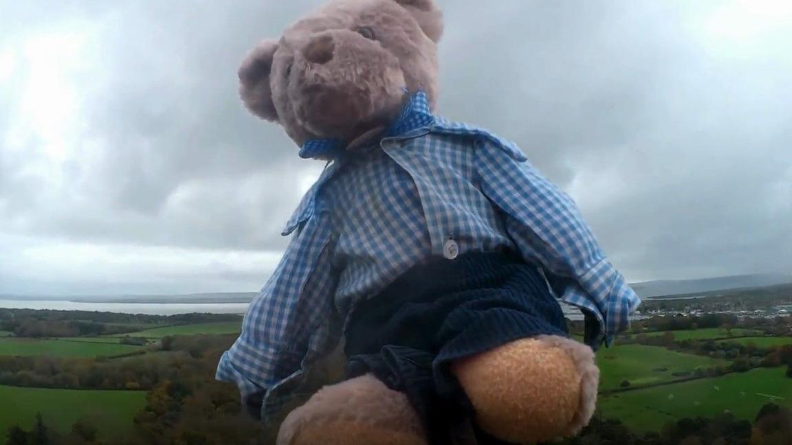 Small, brown bear, wearing school uniform, navy blue shorts and a blue and white checked shirt flying attached to a weather balloon with green fields below.
