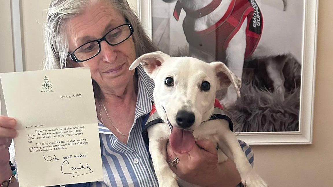 A woman with grey hair and glasses holding up a letter signed from Queen Camilla and a small white Jack Russell with its tongue sticking out