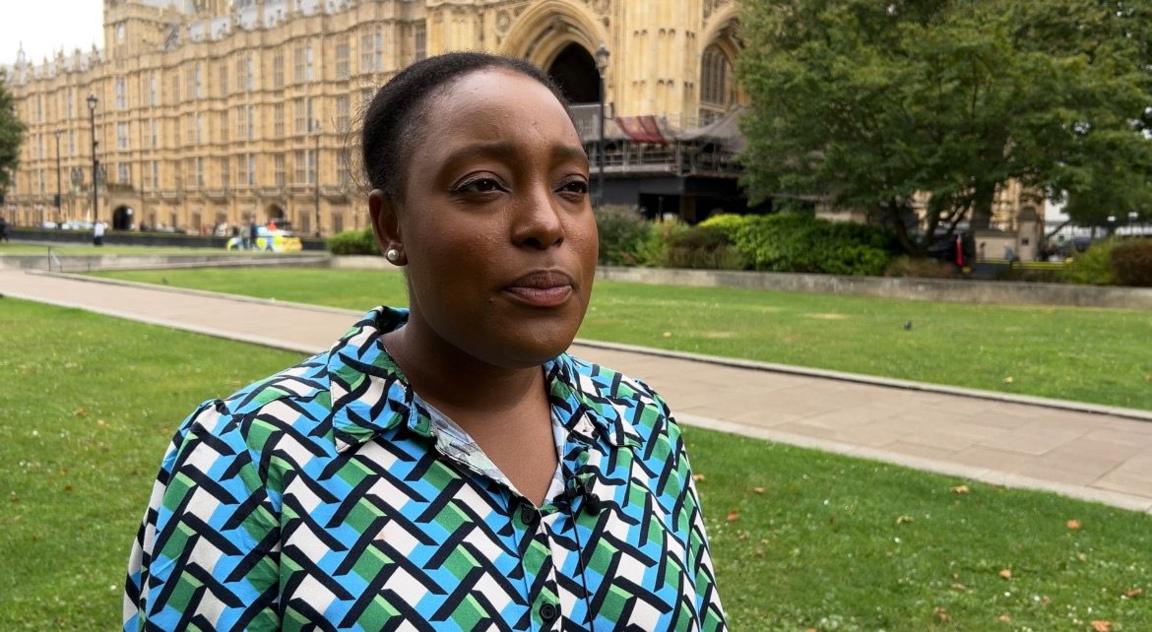 A woman stands on a lawn in front of the houses of parliament. She has black hair and she is wearing a shirt with a blue, green, white and black geometric print.