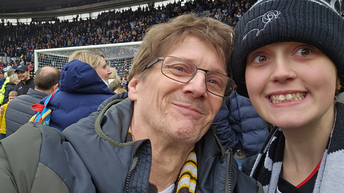 Man with glasses and puffer jacket sitting next to teenage child in bobble hait and a football scarf in a football stadium during a football match