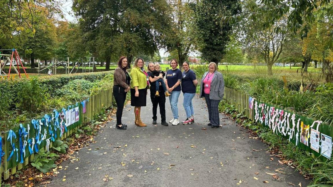 A group of women and young boy posing to the camera along a footpath with blue and pink ribbons on railings either side of them in a park