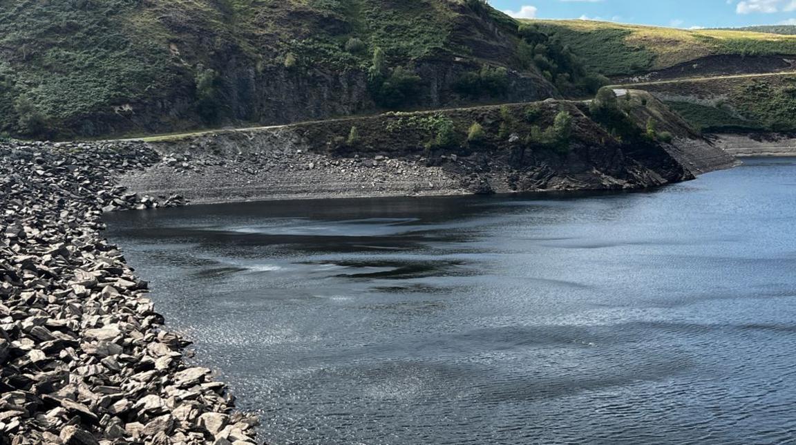 A view from the stone pebble shore at Llyn Brianne. Across the water, the banks of the lake can be seen with horizontal bands of stone emerging out of the water. Beyond the stony banks are hills with grass on them. 