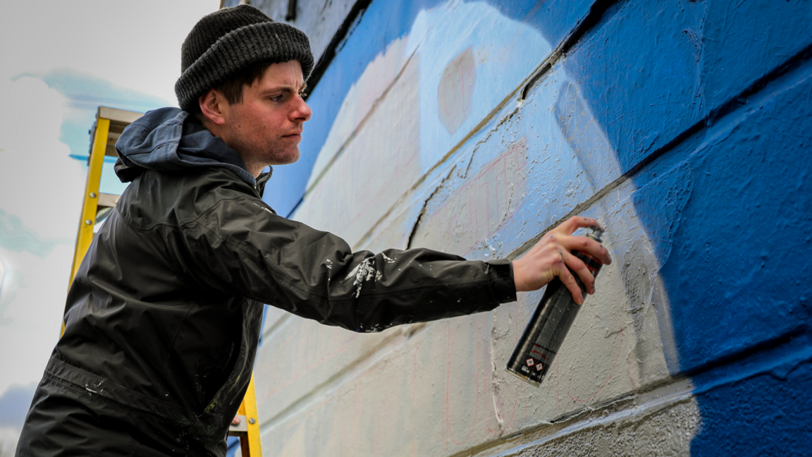 Angry Dan wearing a jacket and woollen hat spraying paint on a wall