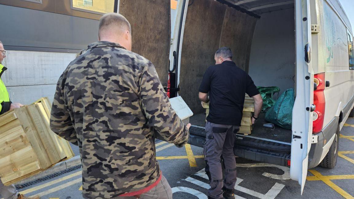 The backs of two men who are unloading hedgehog houses and bird boxes from a white transit van