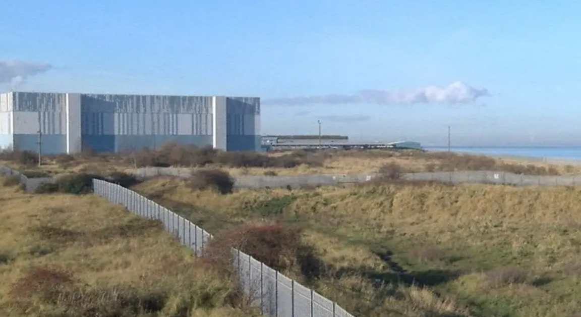 The proposed site is has a large grey building to the left of the shot and is fenced off.  The area is covered in grassland up to the beach.