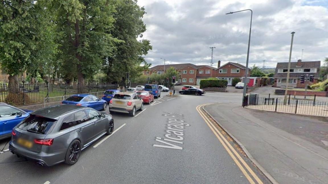 A main road with traffic queued up at a red traffic light at a junction. To the left is a black metal fence with trees behind it. In the background there is a row of red brick buildings