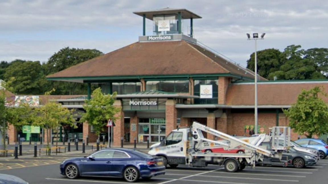 Streetview of several cars parked in a car park with a large supermarket behind. It has the sign saying Morrisons above large glass doors and has a little tower surrounded by brown slanted roofs.