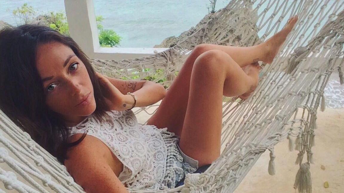 A young woman with long dark hair takes a selfie while sitting in a hammock. The ocean can be seen in the background.