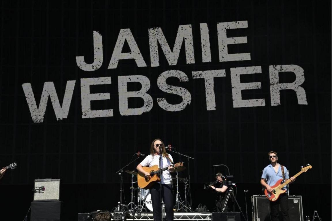 A man onstage with a guitar along side bandmates, with Jamie Webster in letters behind on the backdrop