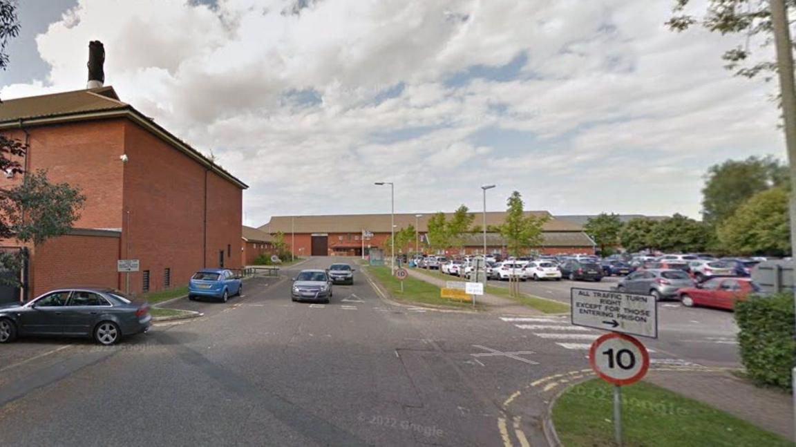 Prison entrance with a large car park and 10mph sign. A single-storey brick building is visible in the background, along with a two-storey brick building to the left. There are some trees in the grounds.