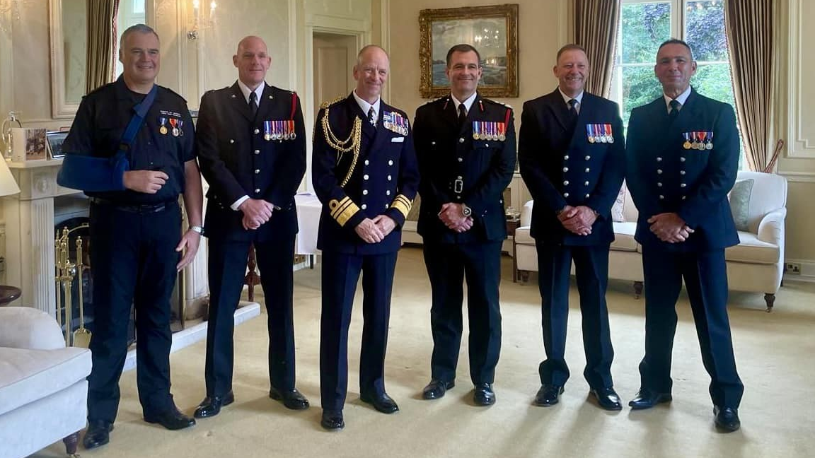 Six men stand in a line wearing navy blue fire service uniforms with about four to seven medals on their left chests. They are in a regency-style cream room with a chandelier, and ornate furniture.