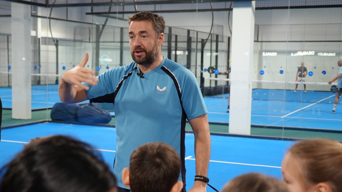 Jason Manford speaking to a group of children while standing on a blue indoor tennis court. He has brown short hair and a brown beard. He is wearing a sky blue T-shirt, the same colour as the court, with black stripes down the sides. As he is speaking, he is gesticulating with his right arm up, elbow bent, his hand raised to chest height and splayed. In the bottom of the image are the top of four children's heads, facing Jason.