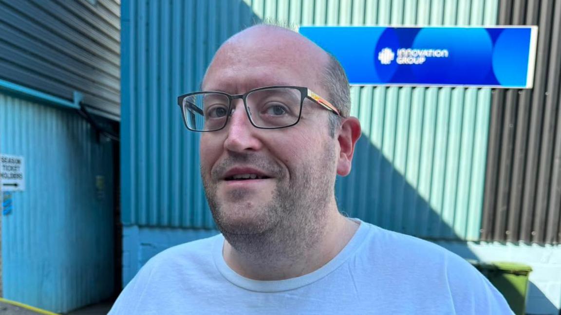 A man smiles for the camera as he stands in front of a blue building, He has black rimmed glassed and stubble.