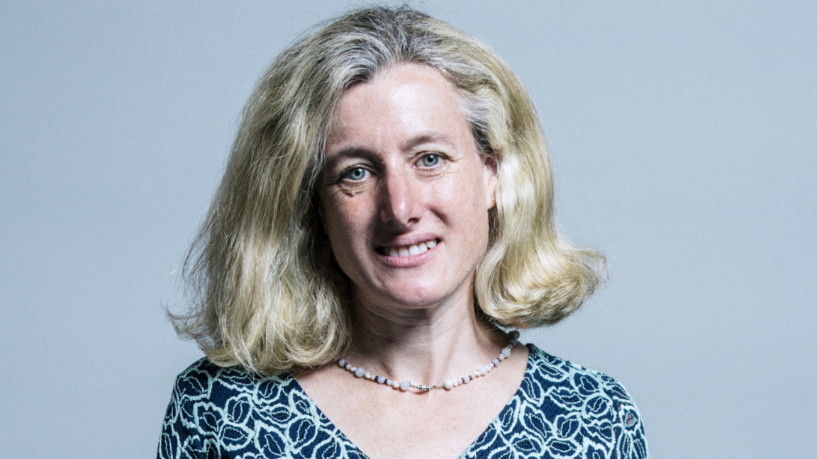 Woman with short blonde hair and a patterned blue top and beaded necklace stands infront of grey background 