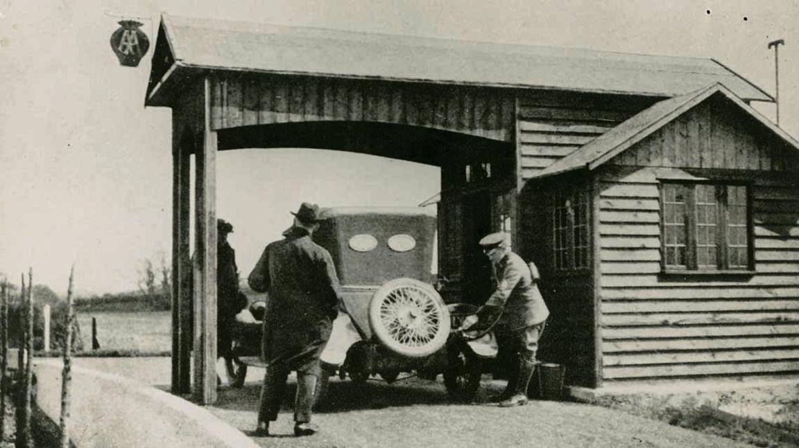 A black-and-white image of the canopied Aldermaston filling station, where a uniformed worker is using a hose to put fuel into a vintage car.