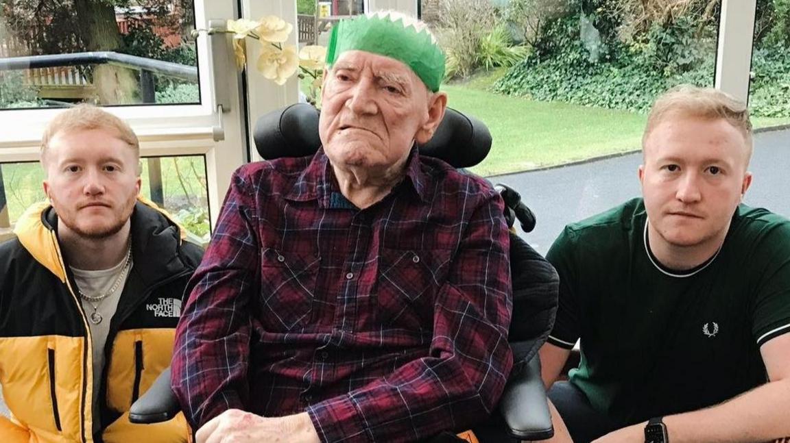 Brian Teasdale sitting in a wheelchair with a headrest, wearing a green paper Christmas party hat and purple and red checked shirt. His twin grandsons - young men  - sit on either side of him. Both have sandy coloured hair. One is wearing a green t-shirt and the other is wearing a black and yellow North Face coat.