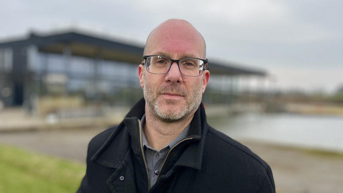 Paul Burrows with the building of St German's pumping station in the background, wearing a black coat, looking at the camera with a neutral expression.