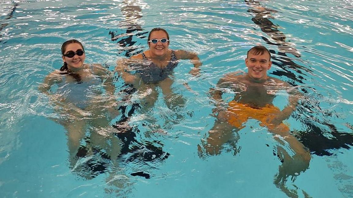 Lois Worrow, Claire Sawyer and James Patient paddling in a swimming pool. They are smiling at the camera. Lois and Claire are wearing goggles.