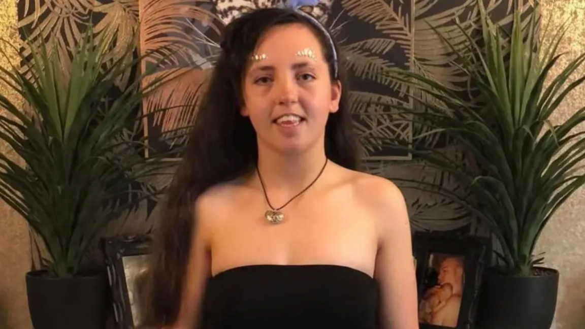 Maria pictured with long black thick hair, wearing a black top, a silver heart necklace and glitter stickers above her eyebrows. There are tow small palm like plants on either side of her.