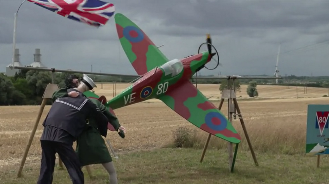 Two life sized scarecrows are kissing, one wearing a sailor uniform, the other wearing a green skirt and jacket. There is a Union Jack flying behind them. A patterned green and burgundy model spitfire plane - with a scarecrow pilot inside - appears to be flying from left to right. In the distance are empty fields trees and grass.