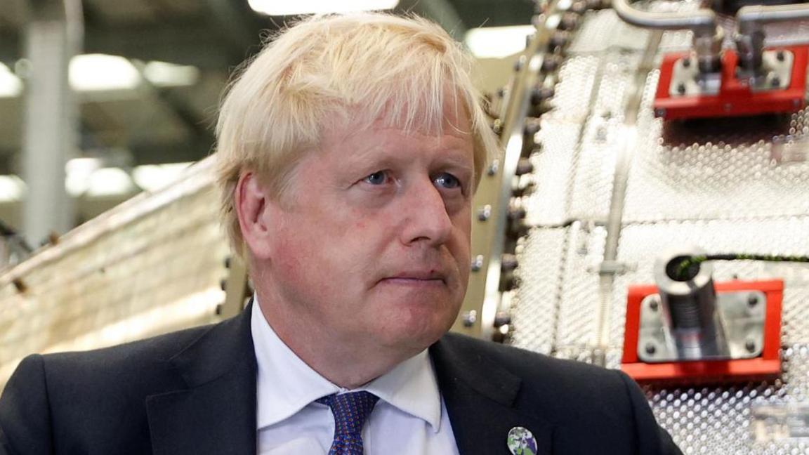 Boris Johnson, with short blonde hair and wearing a black suit, white shirt and blue and red tie, stands in front of industrial equipment