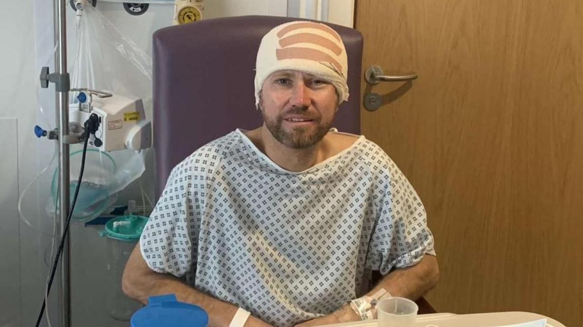 Nick Ball wearing a blue and white patterned hospital gown and a large bandaged around his head. He is sitting in a purple chair in a hospital room, bedside medical equipment and a wooden door. He is looking at the camera but looks weak and unstable.