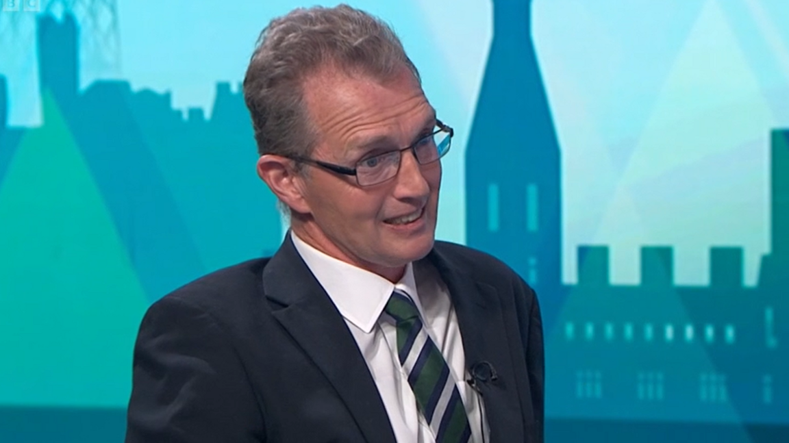 David TC Davies wearing a black suit, white shirt and tie, with blue Politics Wales background