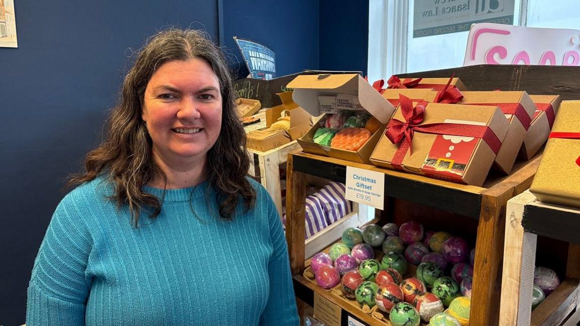 Sarah Prudames with long dark hair smiling at the camera while wearing a blue cardigan with shelves of colourful soaps behind her.